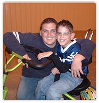 A boy on a Rifton Tricycle posing with a caregiver.