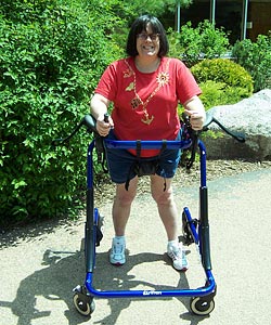 A smiling woman practices gait training outdoors in the sun. She is moving forward in her Rifton gait trainer, which offers more support than any other 4 wheeled walker .