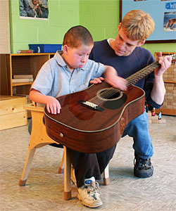 A young boy, in an autism classroom, focuses on strumming a guitar while his teacher patiently frets notes for him.