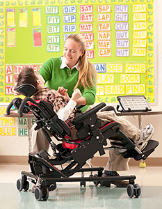 A young boy in a classroom, tilted back in a Rifton adaptive seating chair, smiles at his caregiver who is holding his hand