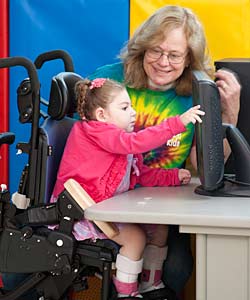 A Young Girl in an Activity Chair Pushes a Screen with the Aide of a RTS Device While a Smiling Woman Assists Her
