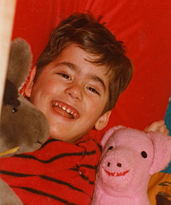 A young Duane lies on a red pillow cuddling with his pink pig and brown horse stuffed animals.