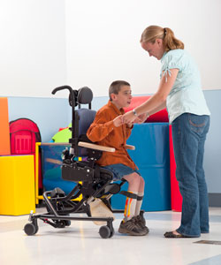A young boy is helped out of his activity chair by a therapist to practice relearning motor skills