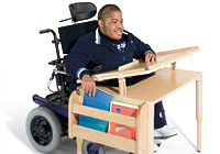 A teenage boy sits in his wheelchair at a desk designed for complex rehab and with multi-use technology