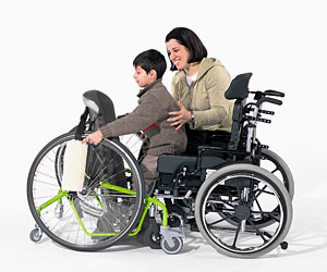 A woman assists a young boy from a wheelchair to an adaptive stander for motor skills training