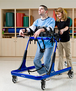 A young man with developmental disabilities is assisted by a therapist to practice walking in a blue Pacer gait trainer 