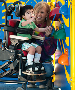 A young boy with Rett Syndrome, grabs with awe at a bell, while being tilted forward in his Rifton Activity Chair by a young woman.