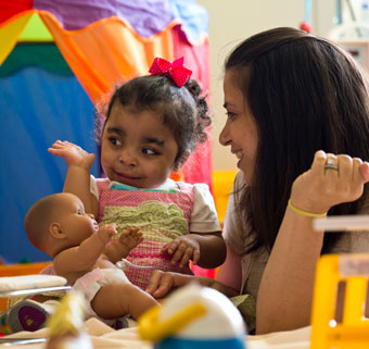 A therapist and daughter in a playroom, both smiling, as the little girl waves while playing with a doll