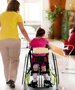 A therapist assists a young girl in a standing device, guiding her through a rehabilitation center