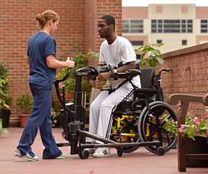 Outside in a courtyard, a therapist uses the Rifton TRAM safe patient handling equipment to transfer a patient from his wheelchair to a standing and mobile position