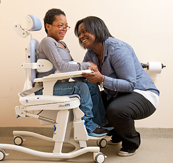 A young girl in a Rifton Hygiene Toileting System smiles at her therapist who knees down in front of her helping her get into proper position