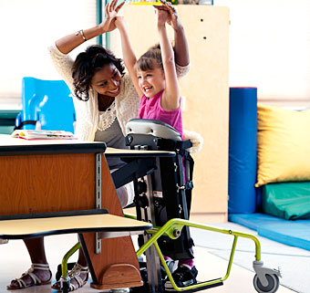 In a classroom setting, a young girl in a Dynamic stander cheers and smiles along with her therapist, raising their arms up in the air. 
