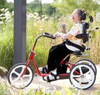 A patient demonstrates equipment positioning while smiling and riding on a red tricycle.