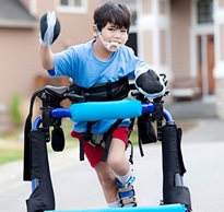 A young boy in an over-ground gait trainer smiles as he walks down the driveway