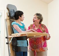 A therapist talks to an adult patient with a neurological condition during a therapeutic standing protocol 