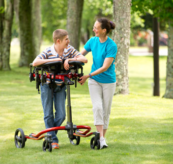 A young boy outside in a park using a dynamic gait trainer is safely guided by his therapist