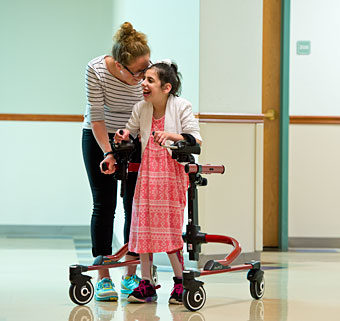 A young girl smiles as she uses a dynamic gait training walker with guidance from her therapist