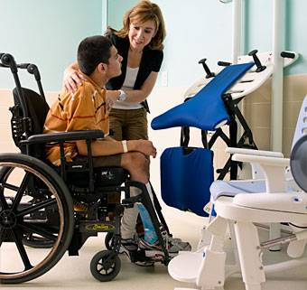 A teacher works with a student to practice motor skills as he does a sit-to-stand transfer from a wheelchair to a support station for toileting 