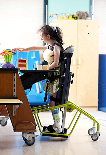 A student at a desk in a classroom uses and adaptive stander to support her while she practices motor skills