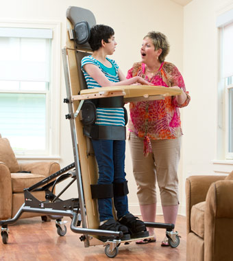 An adult stands in her supine standing device and talks to her therapist.