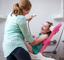 A young girl with special needs smiling in a pink bath chair while her caregiver helps her with this activity of daily living