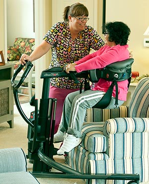 A caregiver in scrubs assists and older lady in a seat to seat transfer using the Rifton TRAM for sphm.