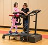 A young girl walks on a treadmill with the aid of a Rifton Pacer, while a caregiver gives her instruction.