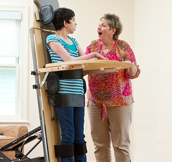 A young woman stands in a Rifton Supine stander, smiling at her caregiver.