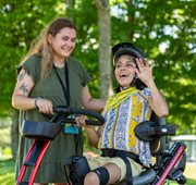 A girl sits on a Rifton Adaptive Tricycle, smiling and waving, with her caretaker next to her.