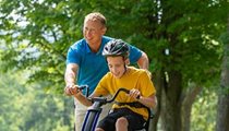 A boy pedals a blue Rifton Adaptive Tricycle, while his therapist walking beside him.