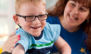 A caregiver smiles at a laughing boy who is standing in a Rifton Prone Stander.