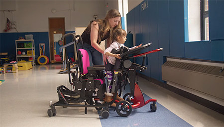 A therapist helps a girl transfer from sitting to standing in a Rifton Stander.