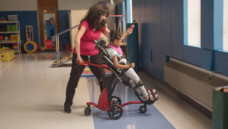 A therapist helps a girl transfer to standing in a Rifton Stander.