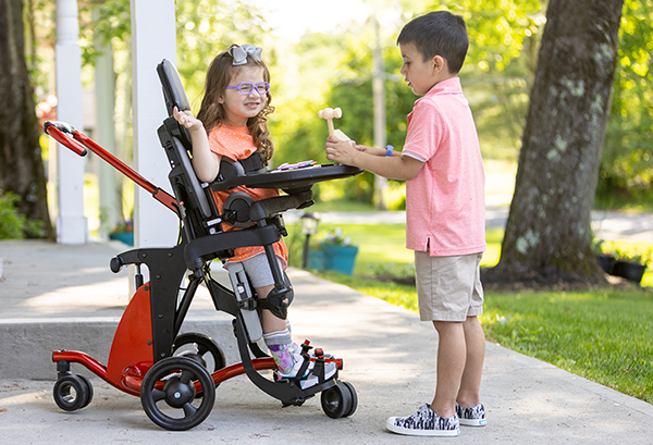 A little girl stands in a Size 1 Rifton Stander and smiles at her brother.