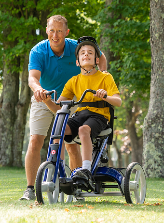A boy rides on a red Rifton Adaptive Tricycle, with his therapist walking beside him.