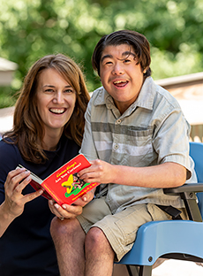 A teenage boy, seated in a Rifton Compass Chair poses for an outdoor photo with his caregiver.