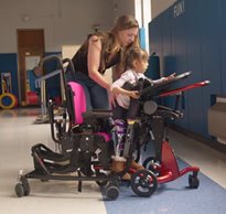 A therapist helps a girl transfer from sitting in a chair to standing in a Rifton Stander.
